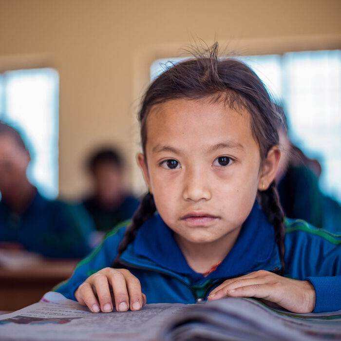 Ein nepalesiches Mädchen liest in der Schule in einem Heft. Im Hintergrund sind weitere Schüler*innen zu sehen.