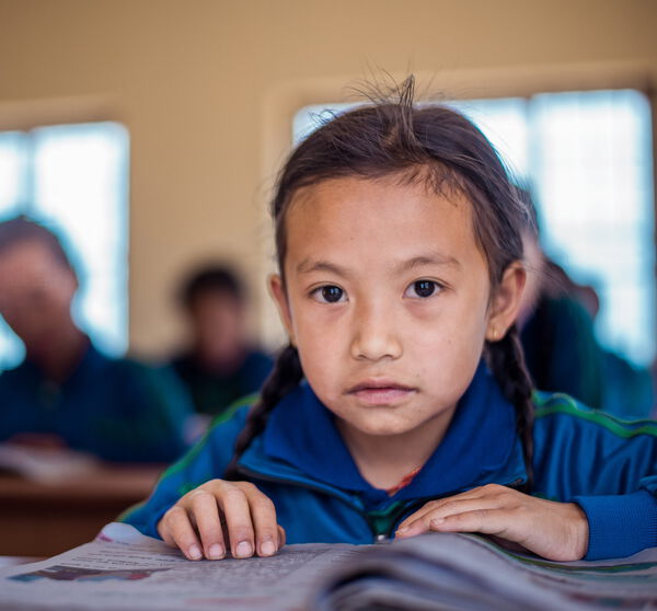 Ein nepalesiches Mädchen liest in der Schule in einem Heft. Im Hintergrund sind weitere Schüler*innen zu sehen.