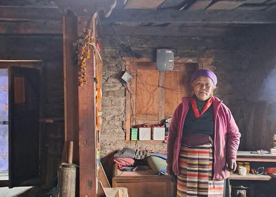 Eine ältere Frau mit einer lila Mütze und einem gestreiften Rock steht in einem schwach beleuchteten Raum mit Wänden aus Ziegeln und Holzbalken. An der Wand hinter ihr ist eine Solarstromanlage befestigt