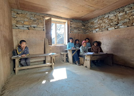 Nepalesiche Kinder sithen im Klassenzimmer afu Holzbänken und folged dem Unterricht.